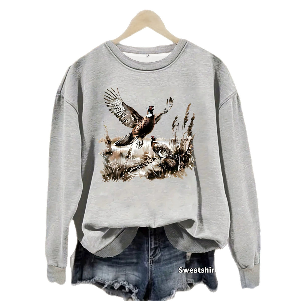 Gray Pheasant Print Crewneck Fleece Sweatshirt on a hanger over denim shorts