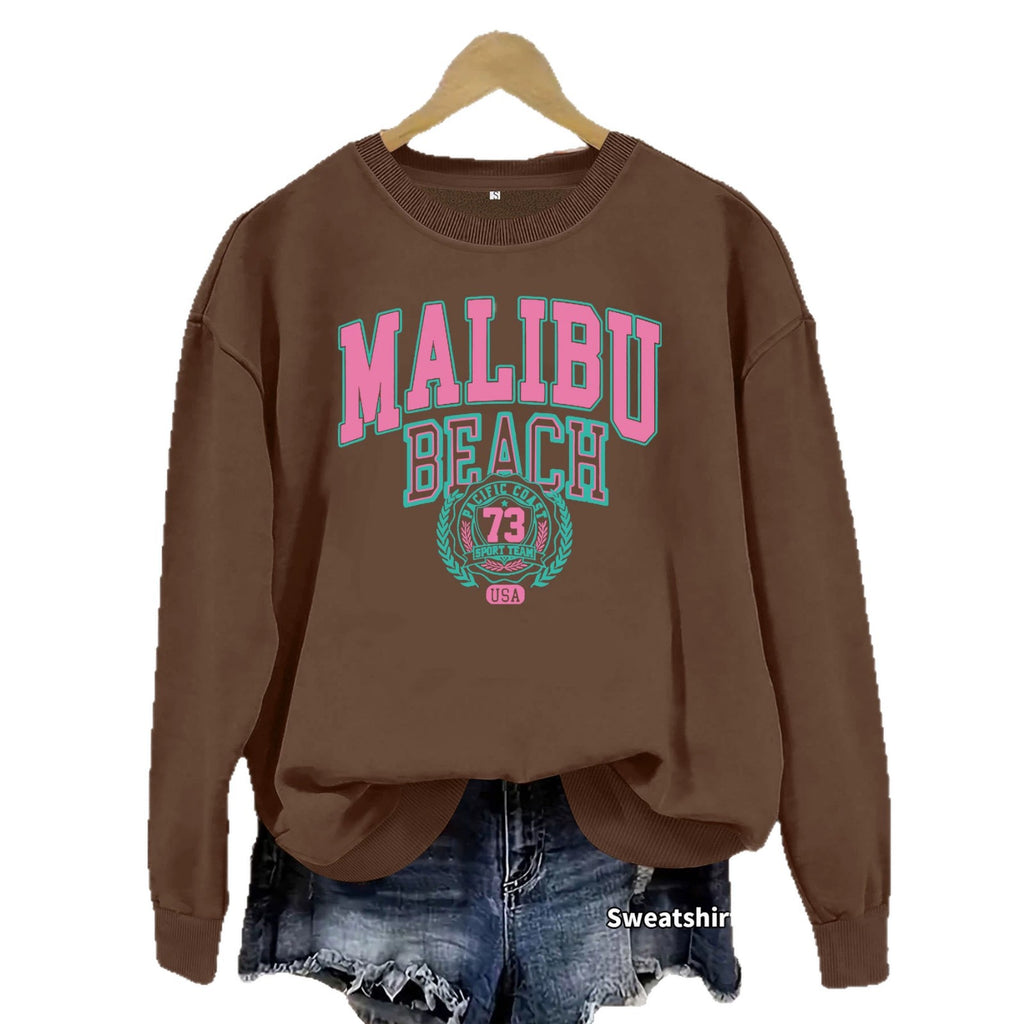 Coffee brown Malibu Beach Graphic Sweatshirt with pink and teal lettering on a hanger over denim shorts