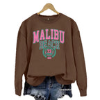 Coffee brown Malibu Beach Graphic Sweatshirt with pink and teal lettering on a hanger over denim shorts