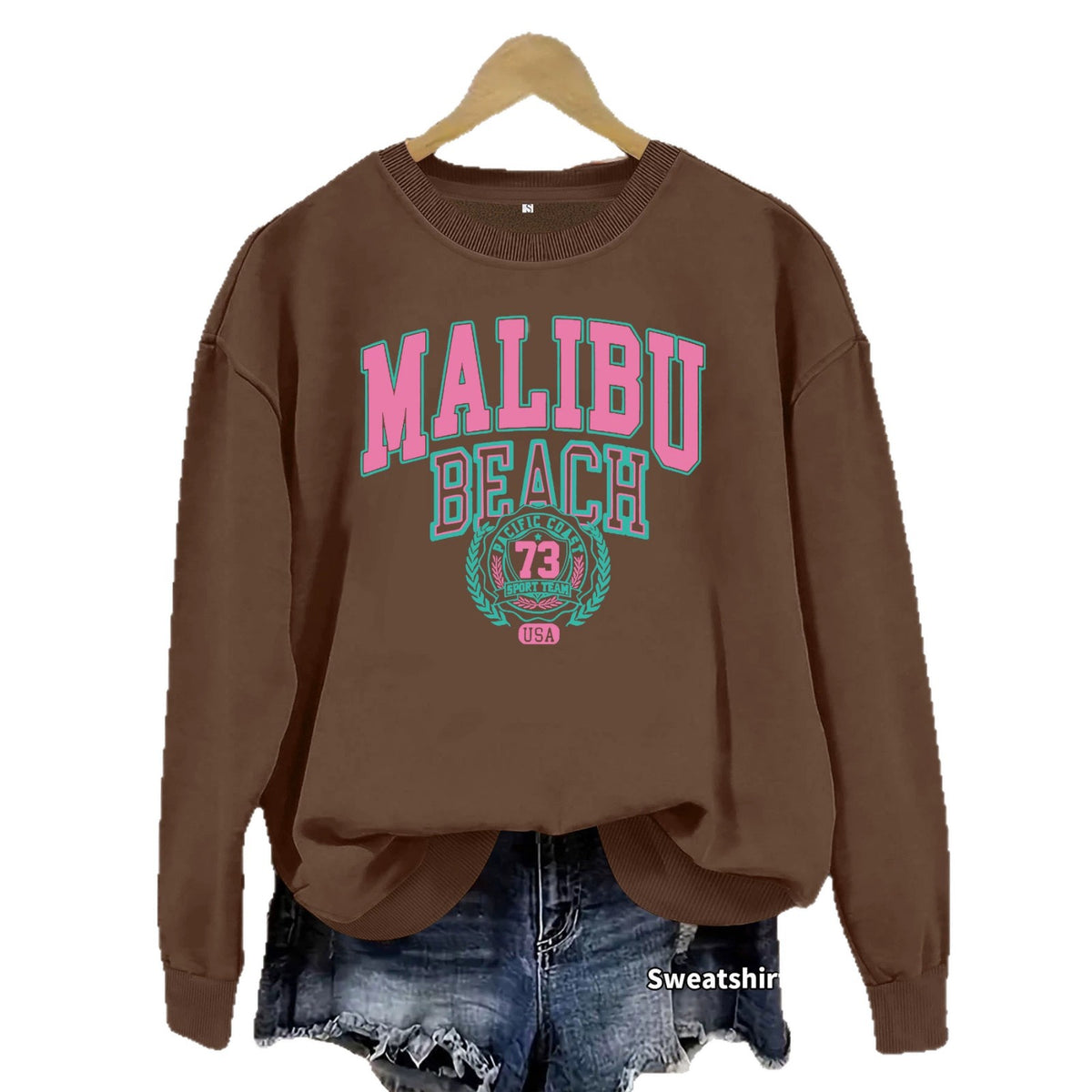 Coffee brown Malibu Beach Graphic Sweatshirt with pink and teal lettering on a hanger over denim shorts